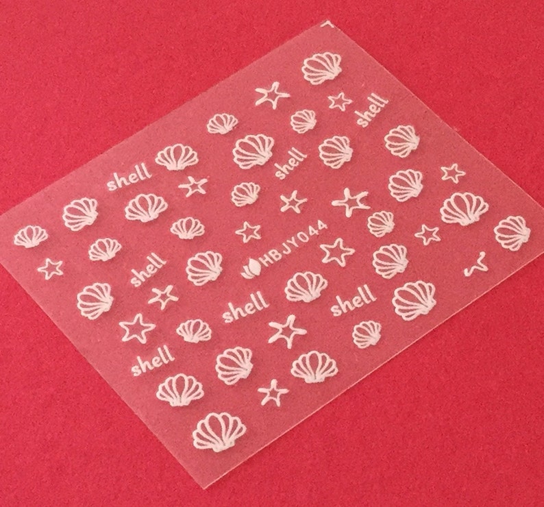 May include: A sheet of white nail decals with images of seashells and starfish. The sheet has the text "HBJY044" and the word "shell" repeated multiple times.