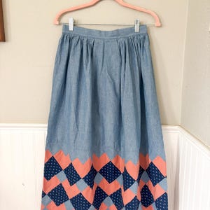 Puede incluir: Falda azul claro con cintura fruncida y dobladillo decorativo. El dobladillo presenta un patrón repetido de formas de diamante coral y azul marino. La falda está colgada en una percha rosa contra una pared blanca.
