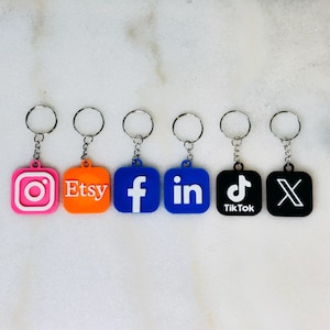 May include: Six keychains with different social media logos. The logos are Instagram, Etsy, Facebook, LinkedIn, TikTok, and X. The keychains are all silver and have a square shape.