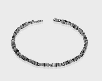 May include: A silver-toned bracelet with a geometric design. The bracelet is composed of small, square-shaped links connected to form a chain. The clasp is visible at one end, and the bracelet is displayed against a plain, light background.