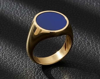 Op de afbeelding: Een gouden zegelring met een diepblauwe ovale middensteen. De ring heeft een gepolijste afwerking en een klassiek ontwerp. De ring wordt getoond op een zwart leren oppervlak.