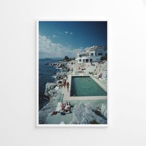 May include: A rectangular swimming pool filled with turquoise water is situated on a rocky cliffside overlooking the dark blue sea. A white multi-story building stands on the cliff above, while people lounge and walk on the concrete pool deck.