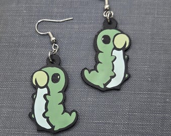 May include: A pair of dangling earrings featuring a cartoon caterpillar design. The earrings have a black outline, with a light green body, white belly, and light yellow head. The earrings are displayed on a dark gray book.