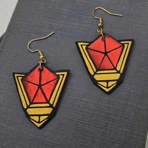 May include: A pair of handmade earrings featuring a red pentagon design within a gold and black shield shape. The earrings have gold-colored hooks and are displayed on a dark gray surface. The earrings are a unique accessory.