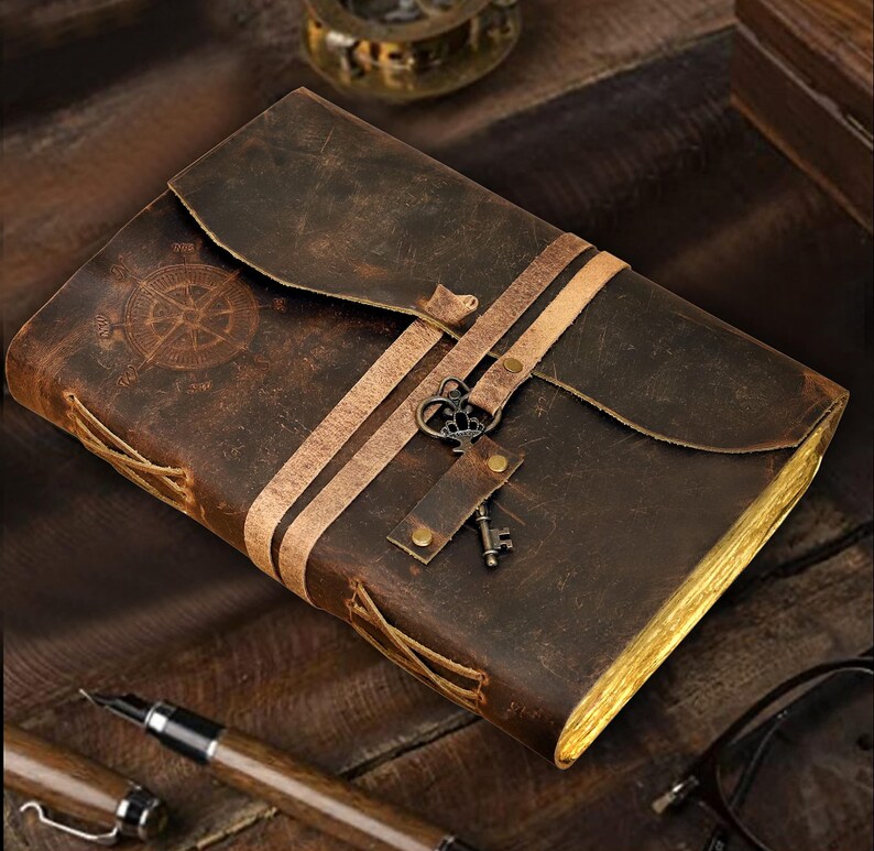 May include: A brown leather-bound journal with a compass design on the front cover. The journal has two leather straps with a decorative metal key attached. The journal is on a wooden surface.