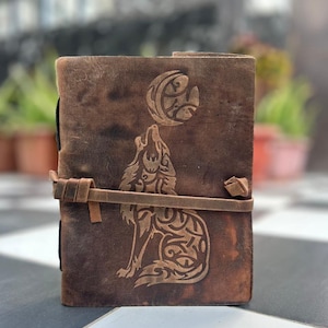 May include: A brown leather journal with a wolf howling at the moon design. The journal has a leather strap closure.