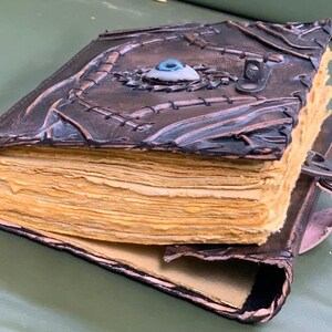 Puede incluir: Un diario de cuero hecho a mano con un dise&ntilde;o &uacute;nico. La cubierta marr&oacute;n oscuro presenta costuras intrincadas y un adorno central en forma de ojo. Las p&aacute;ginas envejecidas son visibles y el diario se asegura con un cierre de metal.