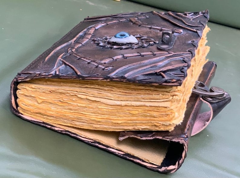 Puede incluir: Un diario de cuero hecho a mano con un dise&ntilde;o &uacute;nico. La cubierta marr&oacute;n oscuro presenta costuras intrincadas y un adorno central en forma de ojo. Las p&aacute;ginas envejecidas son visibles y el diario se asegura con un cierre de metal.