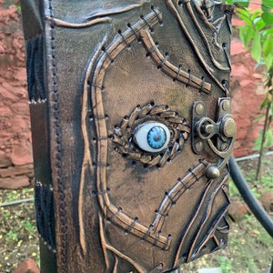 Puede incluir: Un diario de cuero hecho a mano con una apariencia envejecida &uacute;nica. La cubierta presenta un dise&ntilde;o cosido, un cierre de metal y un ojo azul central. El color del diario es una mezcla de marr&oacute;n y bronce, lo que le da una est&eacute;tica vintage.