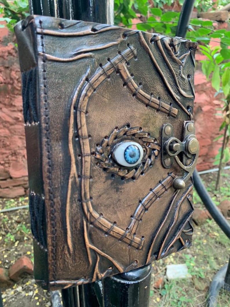 Puede incluir: Un diario de cuero hecho a mano con una apariencia envejecida &uacute;nica. La cubierta presenta un dise&ntilde;o cosido, un cierre de metal y un ojo azul central. El color del diario es una mezcla de marr&oacute;n y bronce, lo que le da una est&eacute;tica vintage.