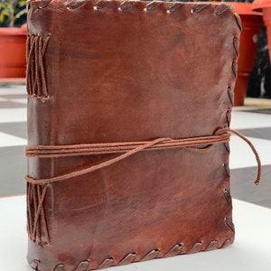 May include: A brown leather journal with a stitched edge and a brown string closure. The journal is closed and lying on a white surface.