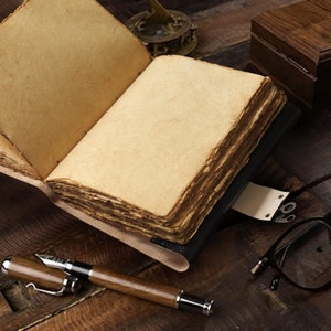 May include: A vintage leather-bound journal with blank pages, open to reveal the aged paper. The journal is lying on a wooden surface with a pair of glasses and a pen nearby.