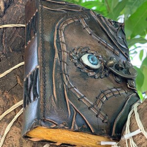 Puede incluir: Un diario de cuero marr&oacute;n oscuro hecho a mano con un dise&ntilde;o de ojo en relieve en la portada. El diario presenta costuras decorativas, un cierre de metal y p&aacute;ginas envejecidas. El ojo tiene un iris azul y una escler&oacute;tica blanca.