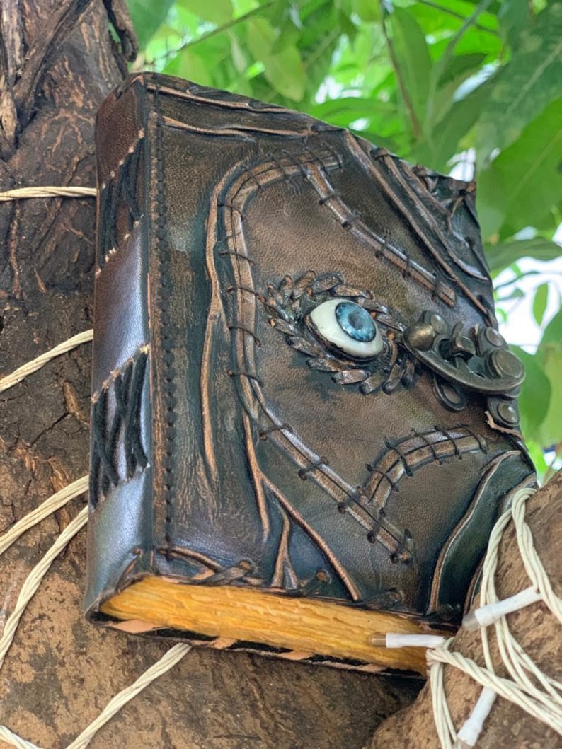 Puede incluir: Un diario de cuero marr&oacute;n oscuro hecho a mano con un dise&ntilde;o de ojo en relieve en la portada. El diario presenta costuras decorativas, un cierre de metal y p&aacute;ginas envejecidas. El ojo tiene un iris azul y una escler&oacute;tica blanca.
