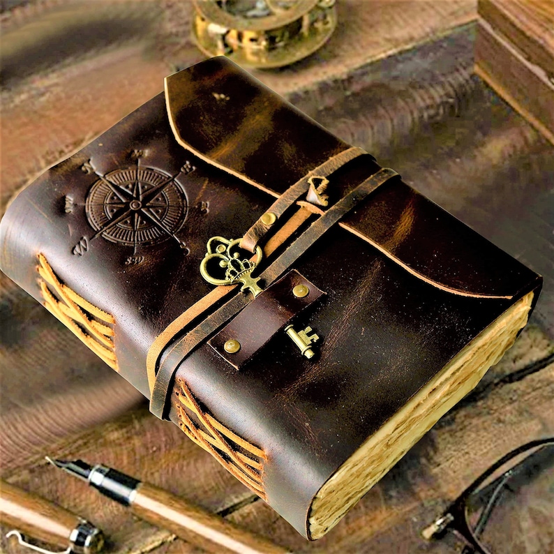 May include: A brown leather journal with a compass design and a key charm. The journal has a rustic look and is tied with leather straps.