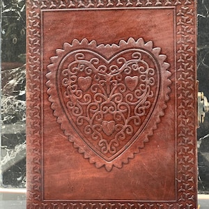 May include: A brown leather journal with a heart-shaped design embossed on the front cover. The heart design features intricate scrollwork and smaller hearts within it.