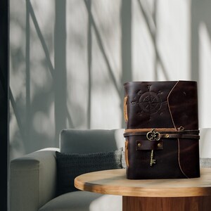 May include: A brown leather-bound journal with a compass design and a key attached to the strap. The journal is sitting on a wooden table in front of a white couch.
