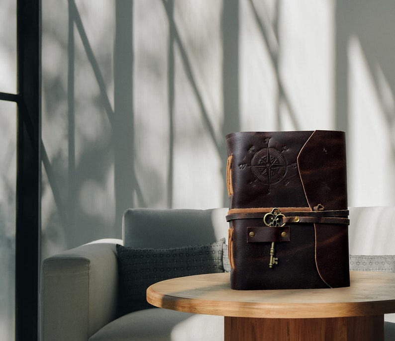 May include: A brown leather-bound journal with a compass design and a key attached to the strap. The journal is sitting on a wooden table in front of a white couch.