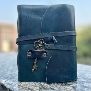 May include: A teal blue leather-bound journal with a decorative key charm and a strap closure.