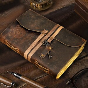May include: A brown leather-bound journal with a compass design on the front cover. The journal has a keyhole and a key attached to it. The journal is closed and lying on a wooden surface.