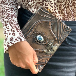 Puede incluir: Un libro de cuero marr&oacute;n con un dise&ntilde;o de ojo azul y cierre met&aacute;lico. El libro tiene una superficie texturizada con detalles cosidos y un ojo decorativo en el centro. El libro est&aacute; en la mano de una persona.