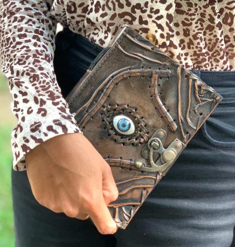 Puede incluir: Un libro de cuero marr&oacute;n con un dise&ntilde;o de ojo azul y cierre met&aacute;lico. El libro tiene una superficie texturizada con detalles cosidos y un ojo decorativo en el centro. El libro est&aacute; en la mano de una persona.