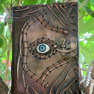 Puede incluir: Un diario de cuero hecho a mano con un dise&ntilde;o &uacute;nico. La cubierta marr&oacute;n presenta detalles cosidos y un patr&oacute;n en relieve. Un ojo azul est&aacute; incrustado en la cubierta y un cierre de metal asegura el diario.