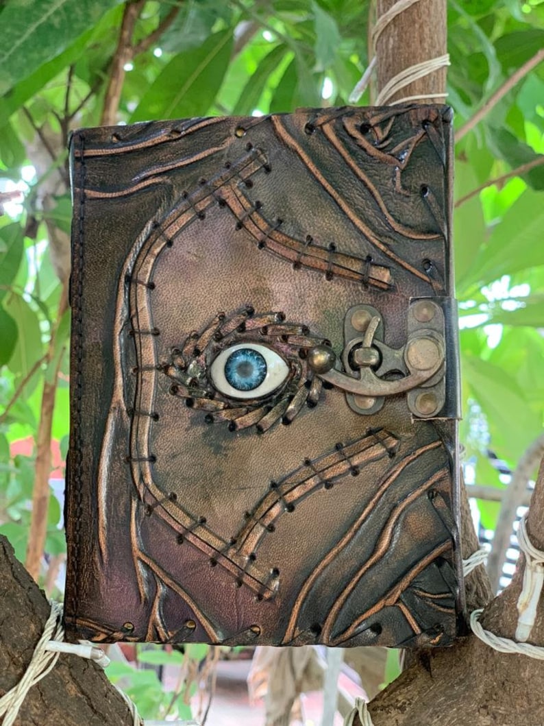 Puede incluir: Un diario de cuero hecho a mano con un dise&ntilde;o &uacute;nico. La cubierta marr&oacute;n presenta detalles cosidos y un patr&oacute;n en relieve. Un ojo azul est&aacute; incrustado en la cubierta y un cierre de metal asegura el diario.