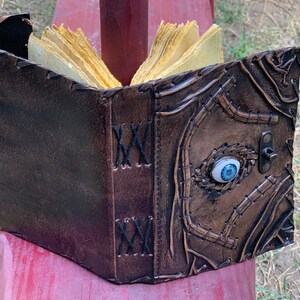 Puede incluir: Un libro artesanal de cuero marr&oacute;n con un dise&ntilde;o &uacute;nico. La cubierta presenta un ojo azul incrustado y costuras intrincadas. Las p&aacute;ginas est&aacute;n amarillentas, d&aacute;ndole una apariencia envejecida. Un cierre de metal asegura el libro.