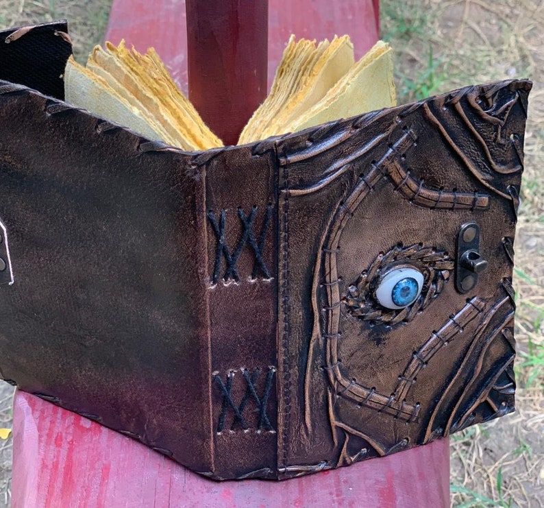 Puede incluir: Un libro artesanal de cuero marr&oacute;n con un dise&ntilde;o &uacute;nico. La cubierta presenta un ojo azul incrustado y costuras intrincadas. Las p&aacute;ginas est&aacute;n amarillentas, d&aacute;ndole una apariencia envejecida. Un cierre de metal asegura el libro.