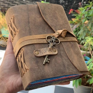 May include: A brown leather journal with a key charm and a strap closure. The journal has a distressed look and is bound with a braided leather cord.