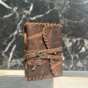 May include: A brown leather journal with a key charm attached to the front cover. The journal has a stitched design and a strap closure.