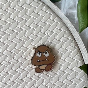 May include: A brown Goomba enamel pin with a gold border, featuring an angry expression. The pin is set against a white, textured background with a woven pattern. The pin is approximately 2.5 cm in height.