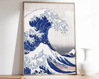 Op de afbeelding: Een ingelijste wandkunstprint met de iconische houtsnede De Grote Golf bij Kanagawa. Het kunstwerk toont een grote, brekende donkerblauwe golf met wit schuim tegen een lichte achtergrond, met een kleine berg zichtbaar in de verte.