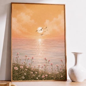 Op de afbeelding: Een ingelijst landschapsschilderij toont een zonsondergang boven de oceaan. Het kunstwerk heeft een warm kleurenpalet met tinten oranje, roze en blauw. Silhouetten van vogels vliegen over de lucht boven het water en roze bloemen staan op de voorgrond.