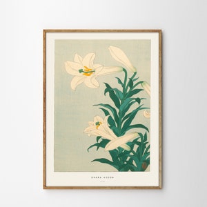 May include: A vintage Japanese woodblock print of white lilies with green leaves against a light blue background. The print is framed in a light brown wood frame. The artist's name, "Ohara Koson", is printed in the bottom right corner of the print.