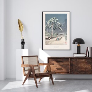 May include: A black framed print of a Japanese woodblock print depicting a snowy scene with a pine tree and figures walking on a path. The print is titled "Sado Garami" and is hanging on a white wall above a wooden dresser.