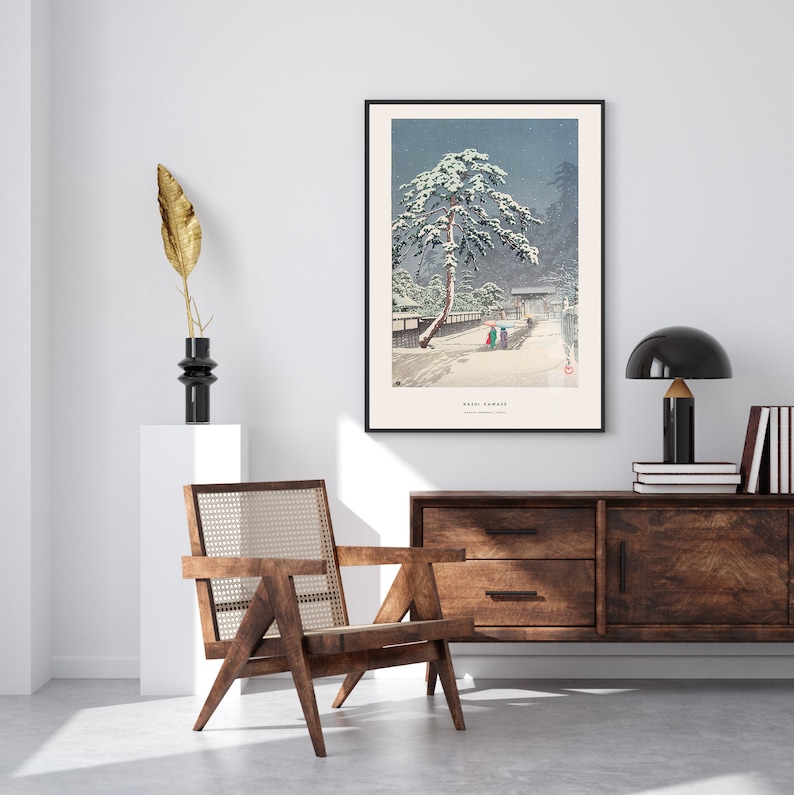 May include: A black framed print of a Japanese woodblock print depicting a snowy scene with a pine tree and figures walking on a path. The print is titled "Sado Garami" and is hanging on a white wall above a wooden dresser.