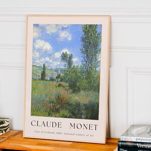 Op de afbeelding: Een ingelijste kunstprint van een schilderij van Claude Monet met de titel "Uitzicht op Vetheuil, 1880." Het schilderij toont een landschap met een veld rode klaprozen op de voorgrond, een dorp in de verte en een blauwe lucht met witte wolken.