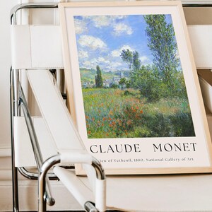 Op de afbeelding: Een ingelijste print van een schilderij van Claude Monet met de titel "Uitzicht op V&eacute;theuil, 1880". Het schilderij toont een landschap met een dorp in de verte, een bloemenveld en een blauwe lucht met witte wolken.