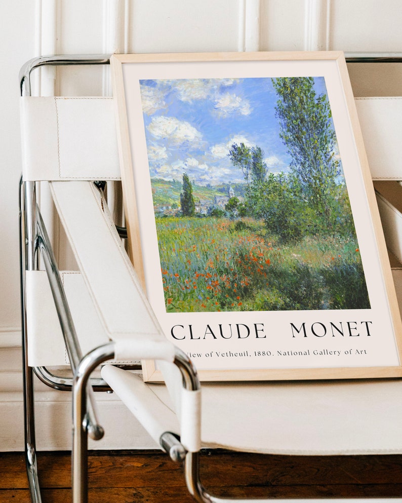 Op de afbeelding: Een ingelijste print van een schilderij van Claude Monet met de titel "Uitzicht op V&eacute;theuil, 1880". Het schilderij toont een landschap met een dorp in de verte, een bloemenveld en een blauwe lucht met witte wolken.