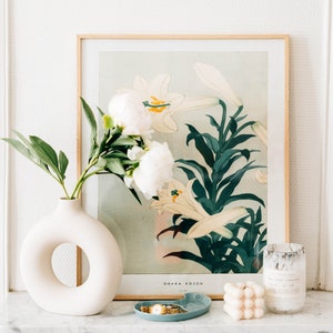 May include: A white ceramic vase with a ring-shaped opening, filled with white peonies and green leaves. The vase is on a white marble surface in front of a framed print of white lilies with green leaves. The print has the text "OMAKA KOSON" and "1918" at the bottom.
