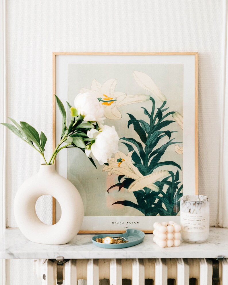 May include: A white ceramic vase with a ring-shaped opening, filled with white peonies and green leaves. The vase is on a white marble surface in front of a framed print of white lilies with green leaves. The print has the text "OMAKA KOSON" and "1918" at the bottom.