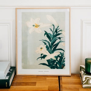 May include: A framed print of a Japanese botanical illustration featuring white lilies with green leaves. The print is titled "Ohara Koson" and is in a light wood frame.