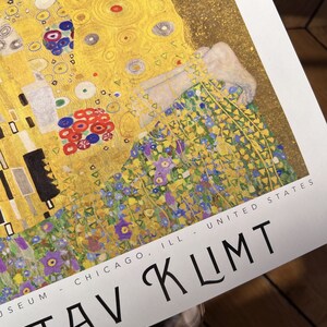 Pu&ograve; includere: Un primo piano di un poster per l'Art Institute di Chicago che presenta un dipinto di Gustav Klimt. Il dipinto &egrave; un ritratto di una donna con uno sfondo dorato e un motivo floreale nella parte inferiore. Il testo "MUSEUM - CHICAGO, ILL - UNITED STATES" e "GUSTAV KLIMT" sono visibili nella parte inferiore del poster.