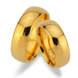 Könnte beinhalten: Zwei goldene Eheringe, einer etwas größer als der andere, mit polierter Oberfläche. Der größere Ring trägt die Inschrift "TREND TRALRINGE" auf der Innenseite.
