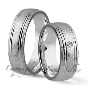 Könnte beinhalten: Ein Paar silberne Trauringe mit gebürsteter Oberfläche. Ein Ring ist mit einem einzelnen Diamanten besetzt.