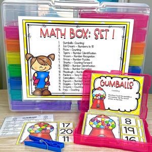 May include: A clear plastic storage box filled with colorful compartments. Inside, a pink box holds cards with gumball machine illustrations and numbers. The main sign reads "MATH BOX: SET 1" with various math activities listed. A blue clothespin is also visible.