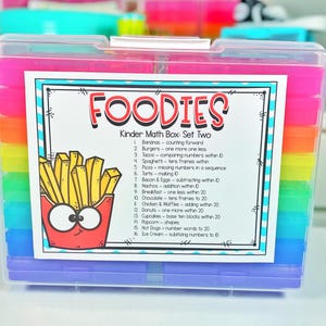 May include: A clear plastic box with a colorful rainbow of compartments. The box is labeled "FOODIES Kinder Math Box: Set Two" and lists various food-themed math activities for kindergarteners.