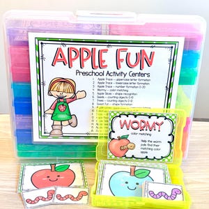 May include: A clear plastic storage case containing colorful smaller containers, with a label titled APPLE FUN Preschool Activity Centers. A smaller yellow container holds cards featuring a red apple, a blue apple, and pink worms.